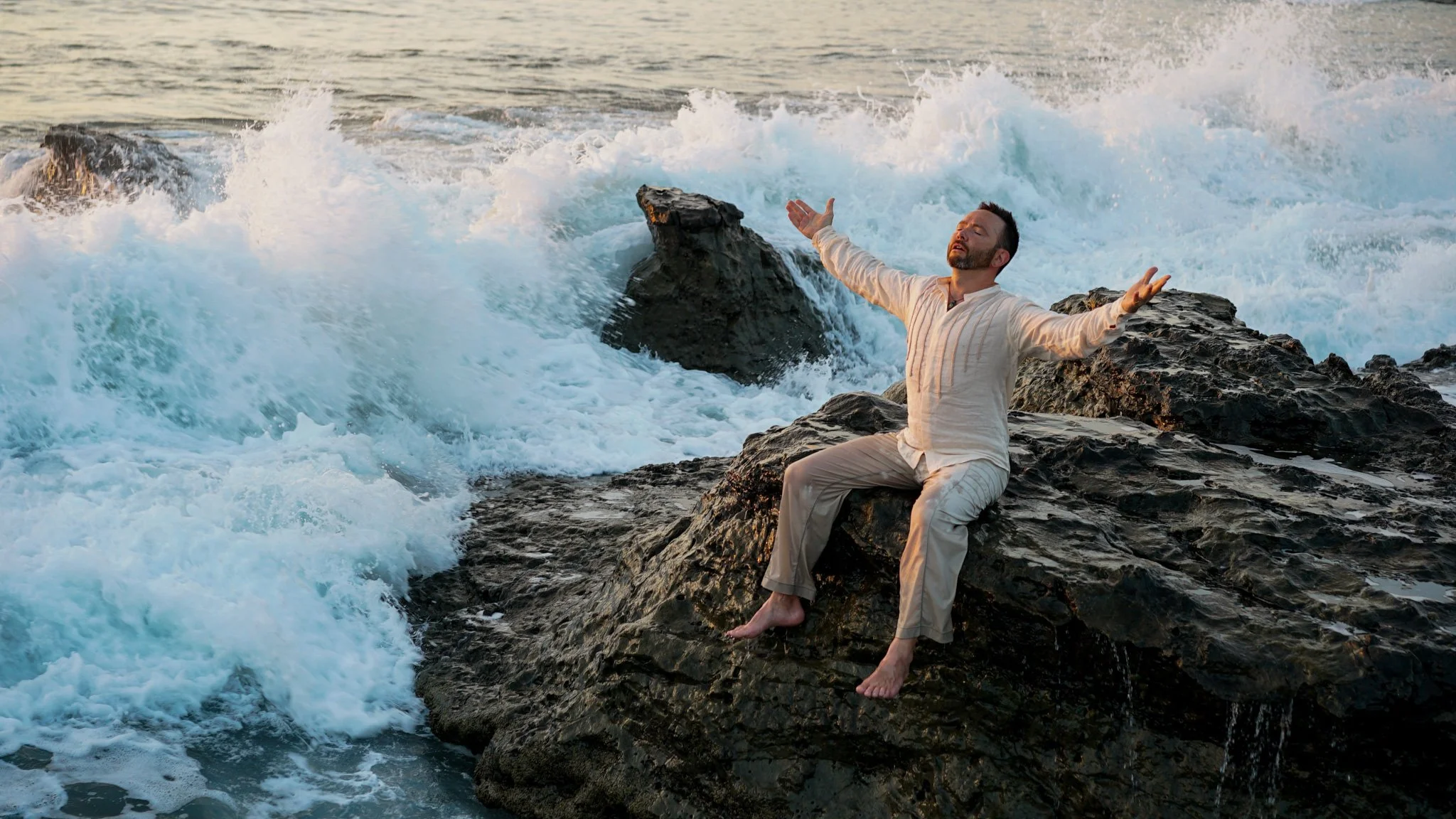 Dr. Dylan Jonlynn on the ocean rocks with arms open to the waves